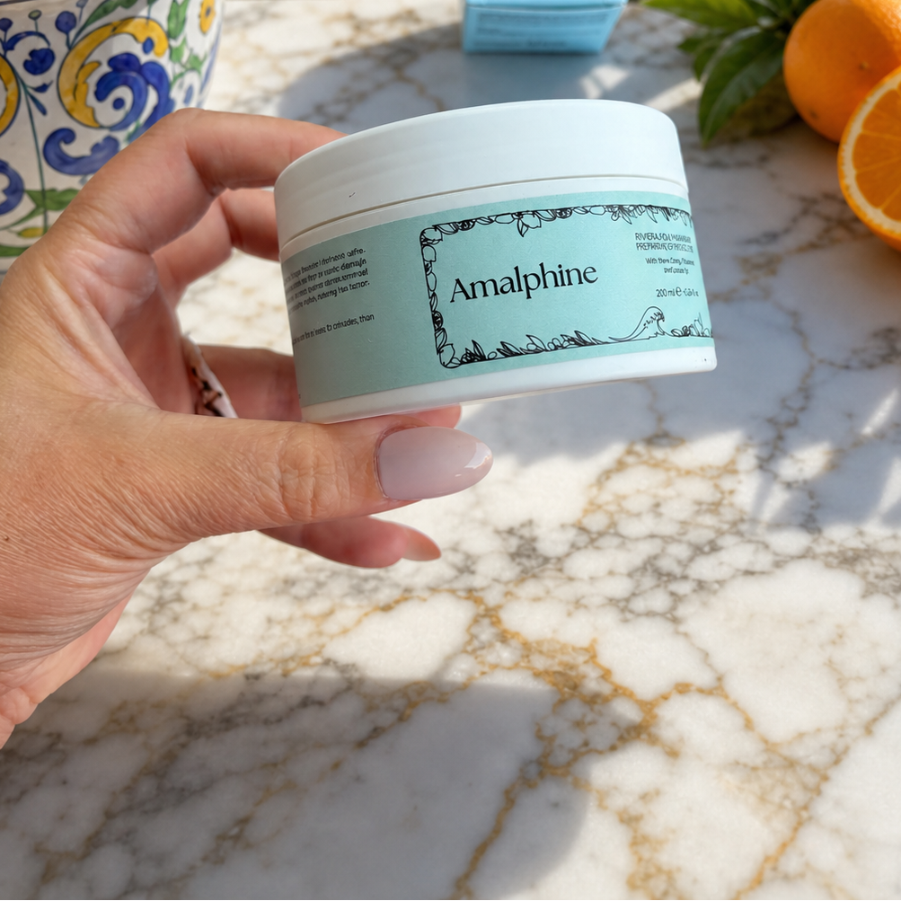 A hand holds a white jar of Amalphine Hair Mask with a mint green label, set against a scenic Mediterranean background with oranges, a patterned bowl, and a blue sea and hillside town.